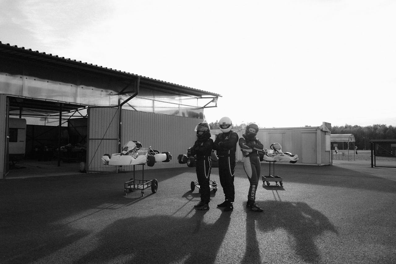 Three go-kart racers in full gear standing with their karts outdoors in monochrome.